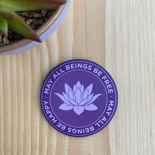 Purple magnet with lotus design and text, may all beings be free may all beings be happy, on a wooden surface.