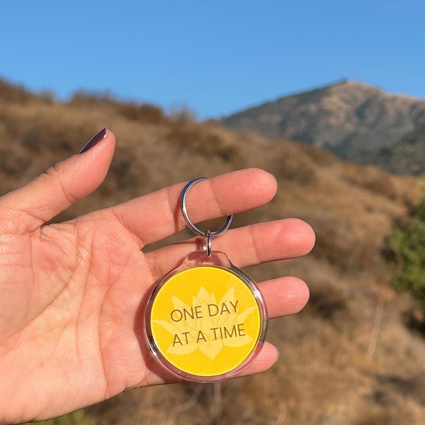 Hand holding a 2" yellow lotus keychain with 'One Day at a Time' text against a mountainous background.
