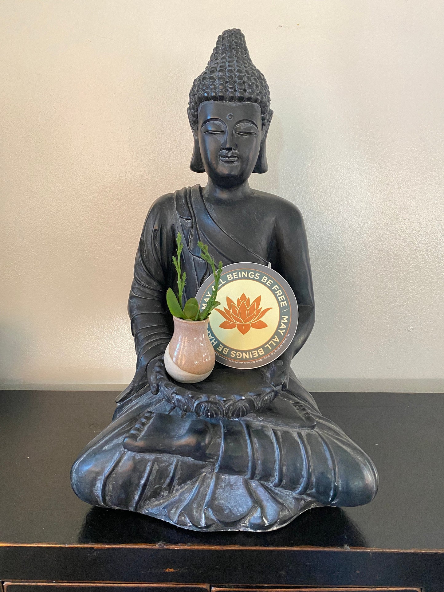 Buddha statue holding a pot with a plant on a plain background with a 4" circular sticker with a saffron colored lotus flower design and Mettā prayer.