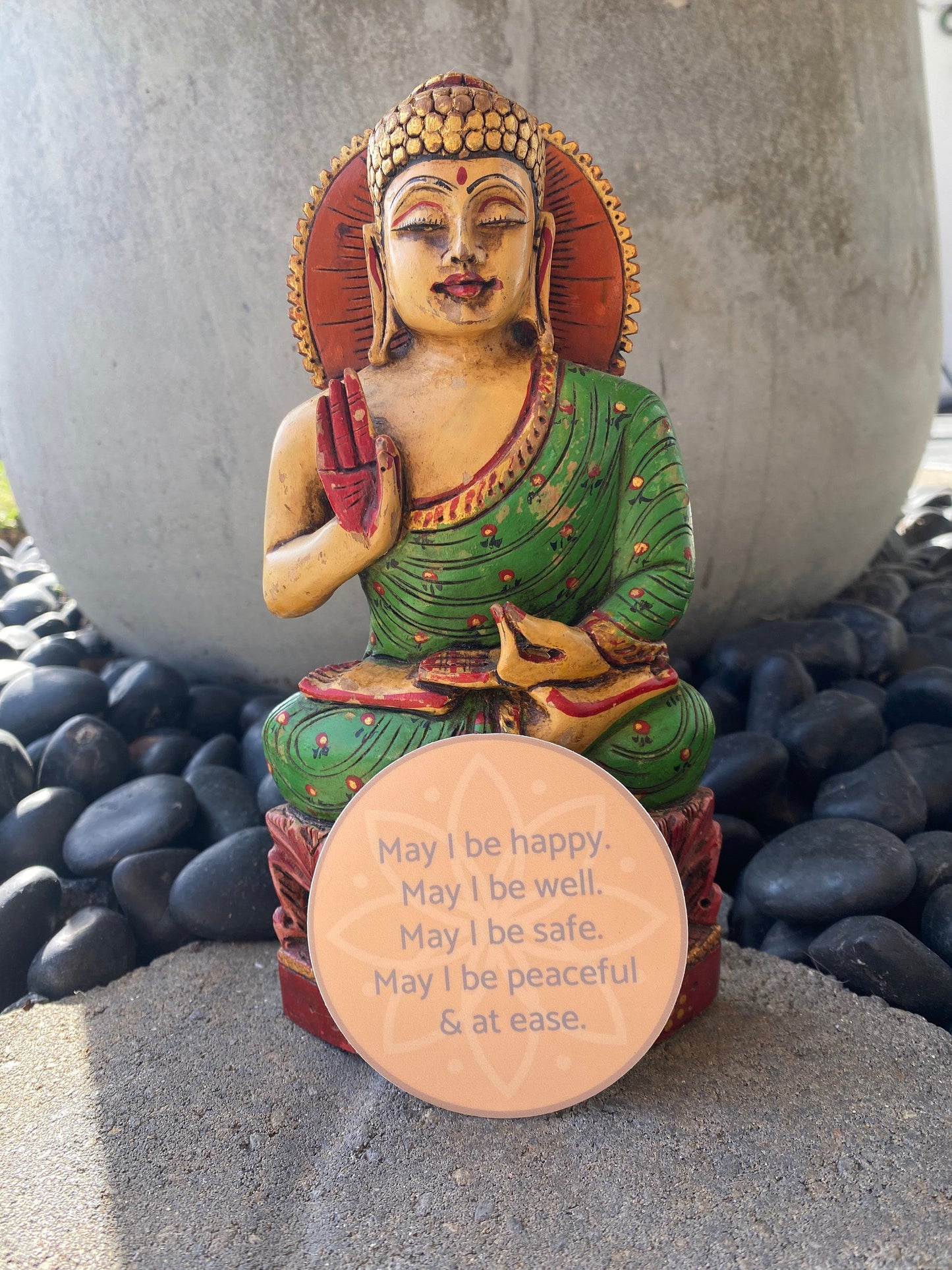 Colorful Buddha statue with a circular sticker with text, may i be happy may i be well may i be safe may i be peaceful may i be at ease, on pebbles.