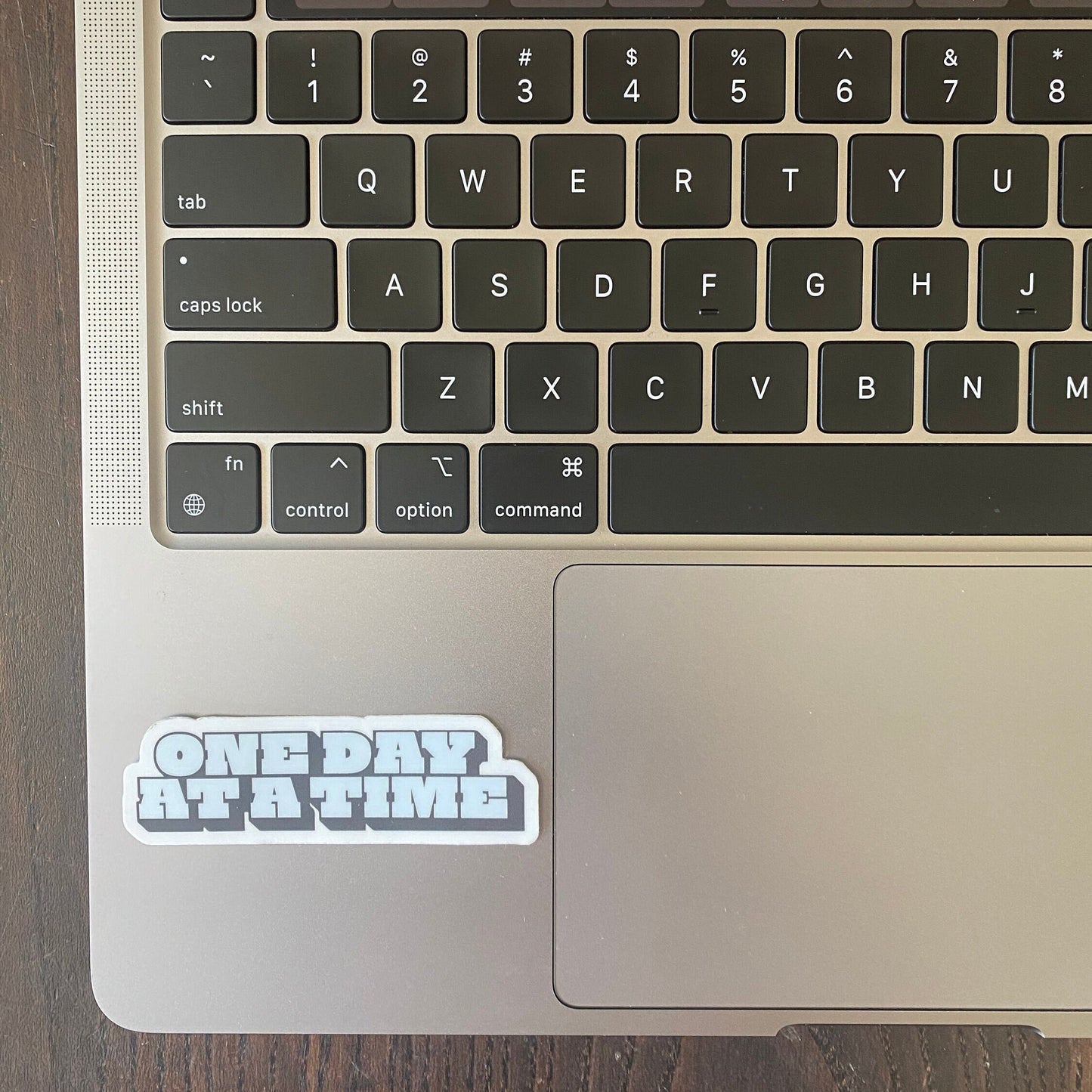 Laptop keyboard with a 'ONE DAY AT A TIME' sticker on a wooden surface