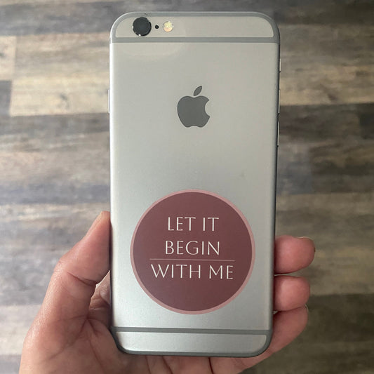 iphone with a pink 2" round sticker with the text held, 'Let It Begin With Me,' in a hand against a wooden background.