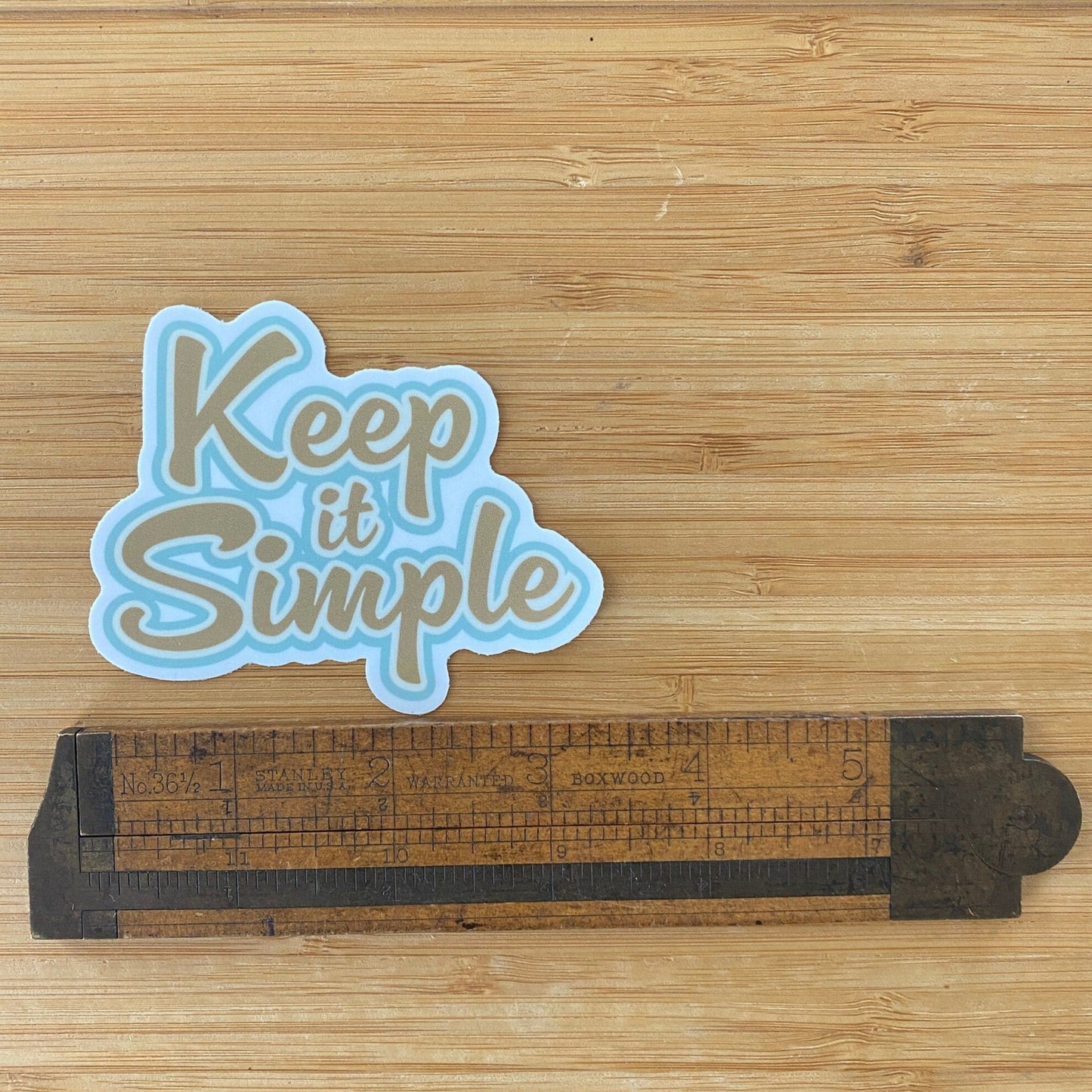 12 Step recovery slogan sticker in gold and blue with the text, 'Keep it Simple' in front of green foliage. on a wooden surface with a ruler for scale.