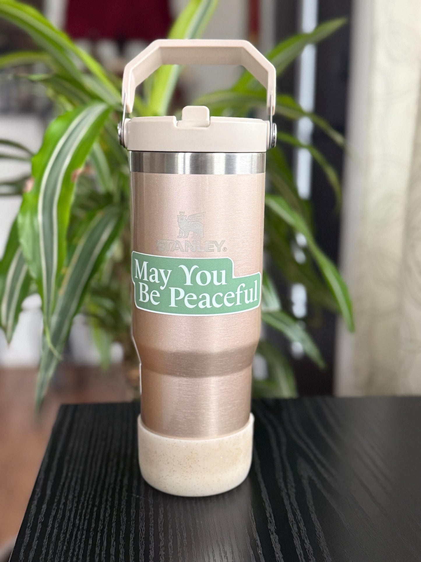 Stanley rose gold tumbler with 'May You Be Peaceful' sticker on a black surface with a plant in the background.