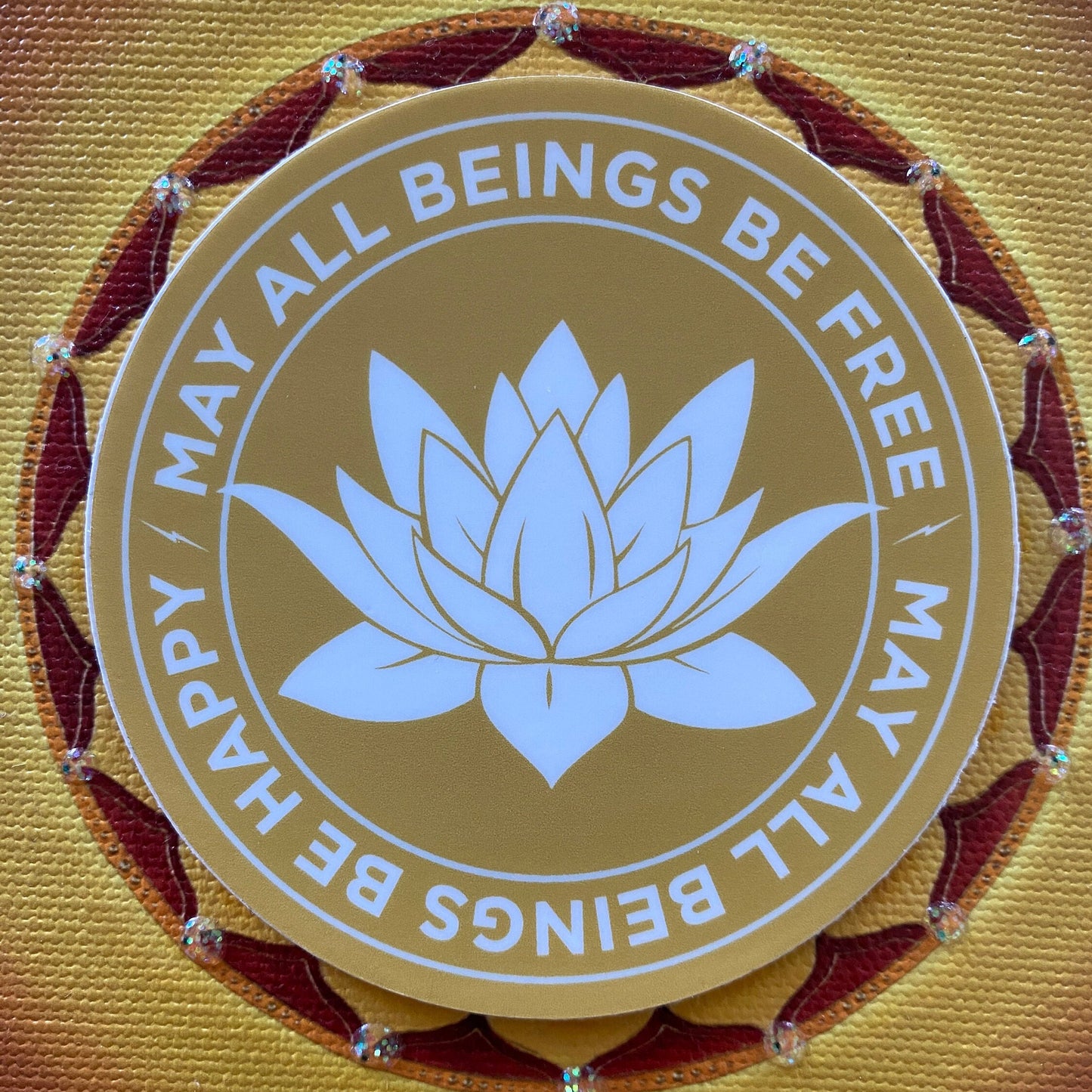 3" gold and white sticker with a lotus flower design and text, may all beings be free may all beings be happy, on a yellow background