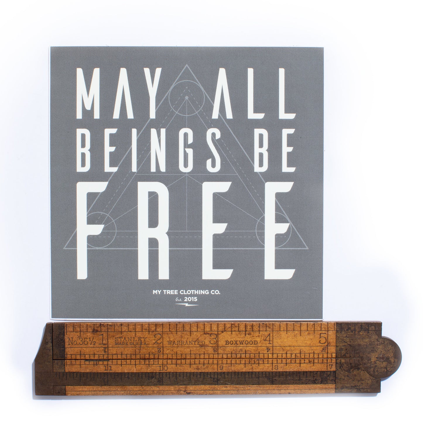 5" square grey sticker with 'May all beings be free' text with sacred geometry design being measured by an antique ruler.
