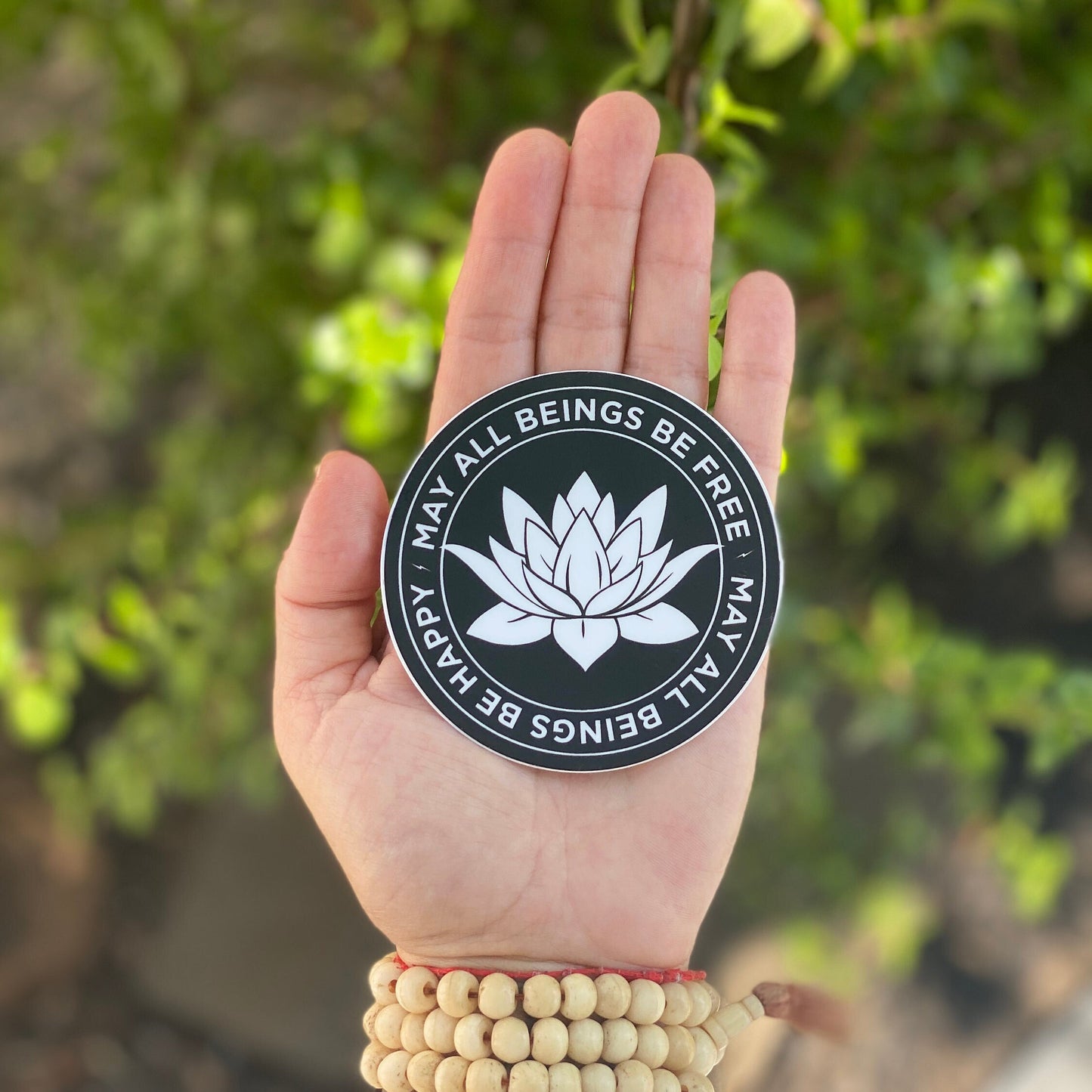 Hand holding a black and white 3" circular sticker with a white lotus flower design and text, may all beings be free may all beings be happy, against a blurred green background.