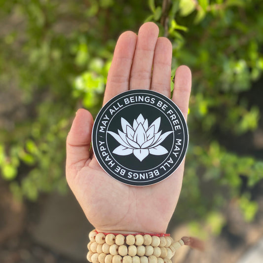 Hand holding a black and white 3" circular sticker with a white lotus flower design and text, may all beings be free may all beings be happy, against a blurred green background.
