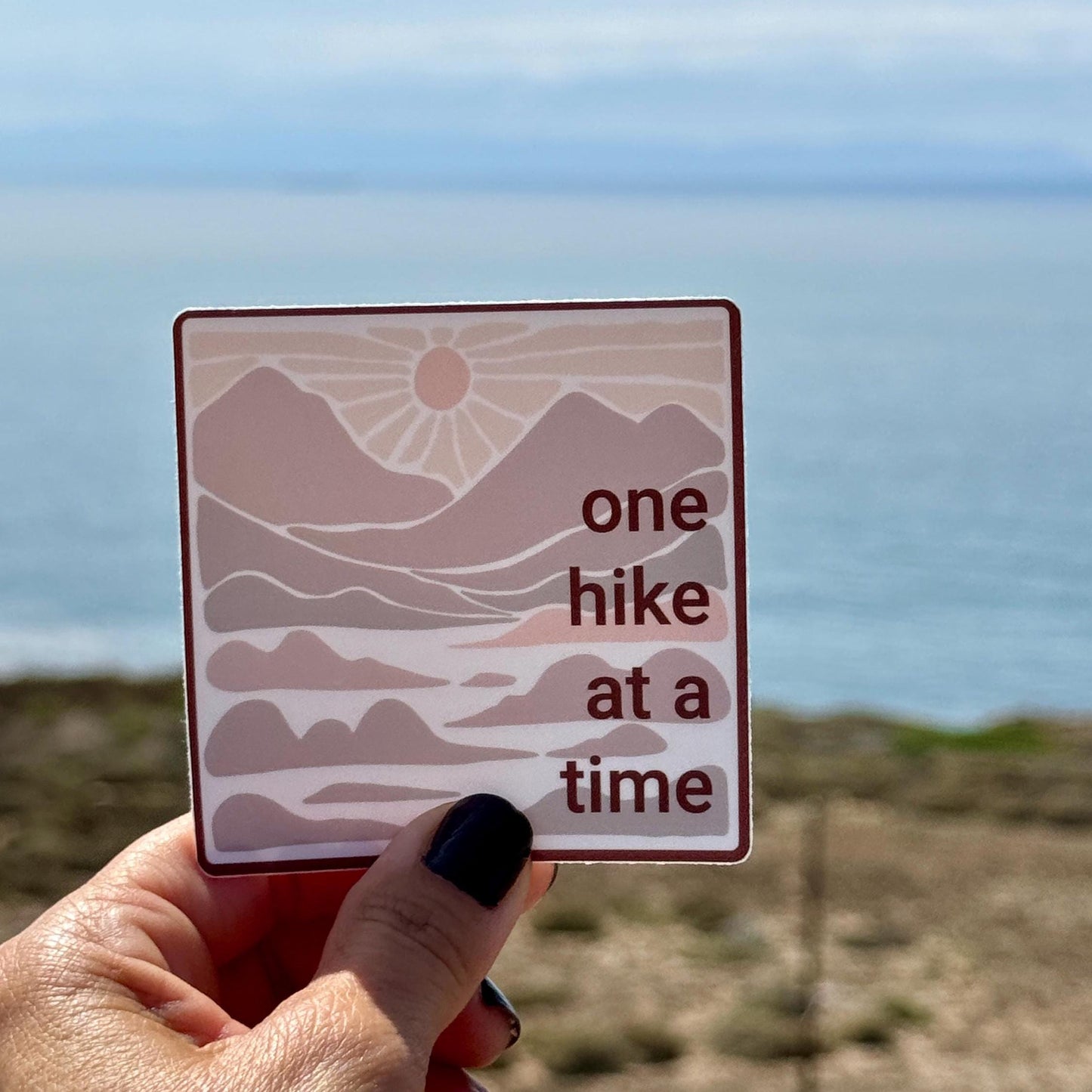 3" square sticker with 'one hike at a time' in neutral colors with mountains and a sunset against a scenic ocean background.