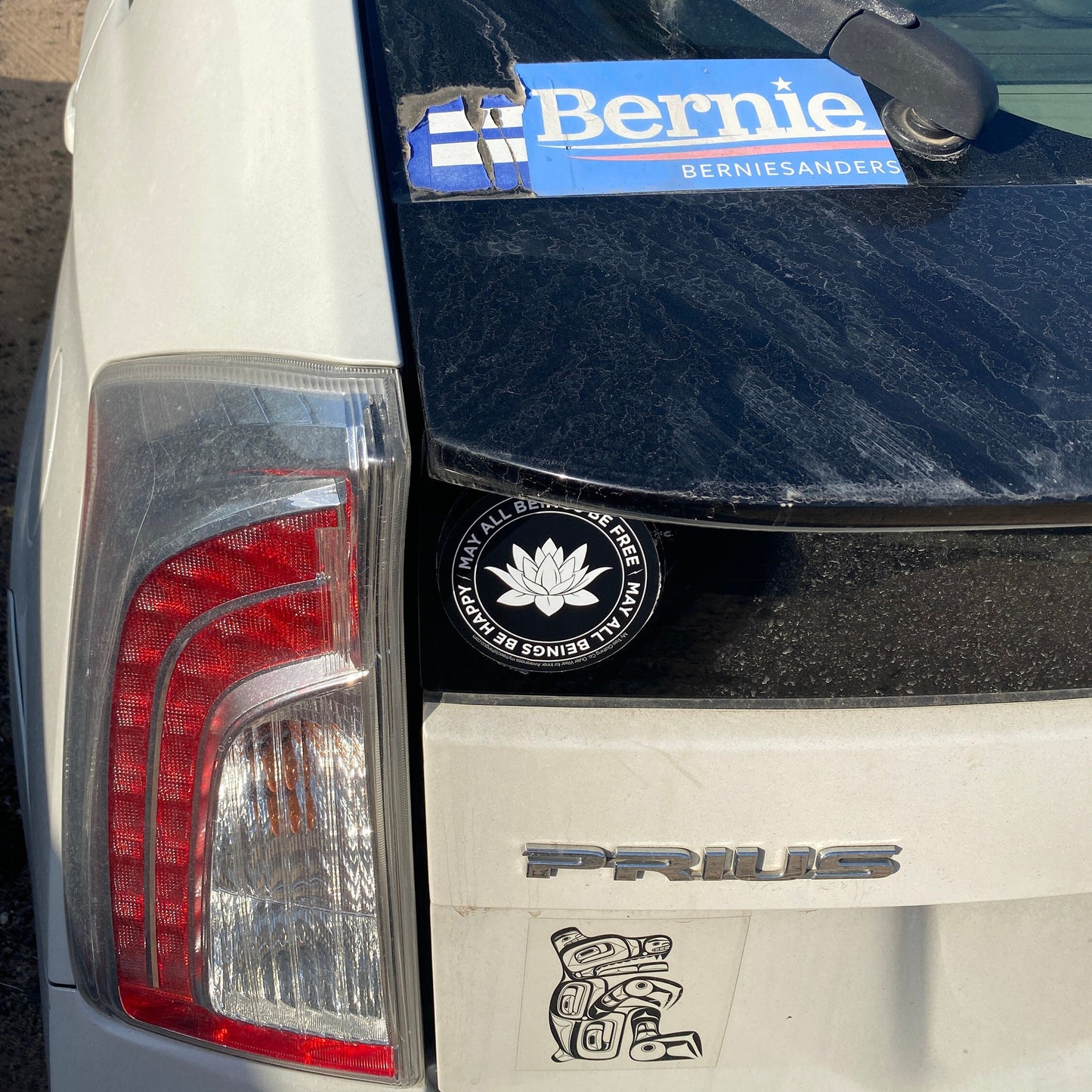 Back of a white Prius with 'Bernie' Sanders sticker and a black and white lotus sticker with the mettā prayer, MAY ALL BEINGS BE FREE MAY ALL BEINGS BE HAPPY.