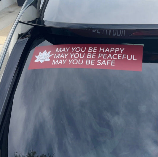 Red and white bumper sticker with a lotus and the prayer, may you be happy may you be peaceful may you be safe, on a car window.