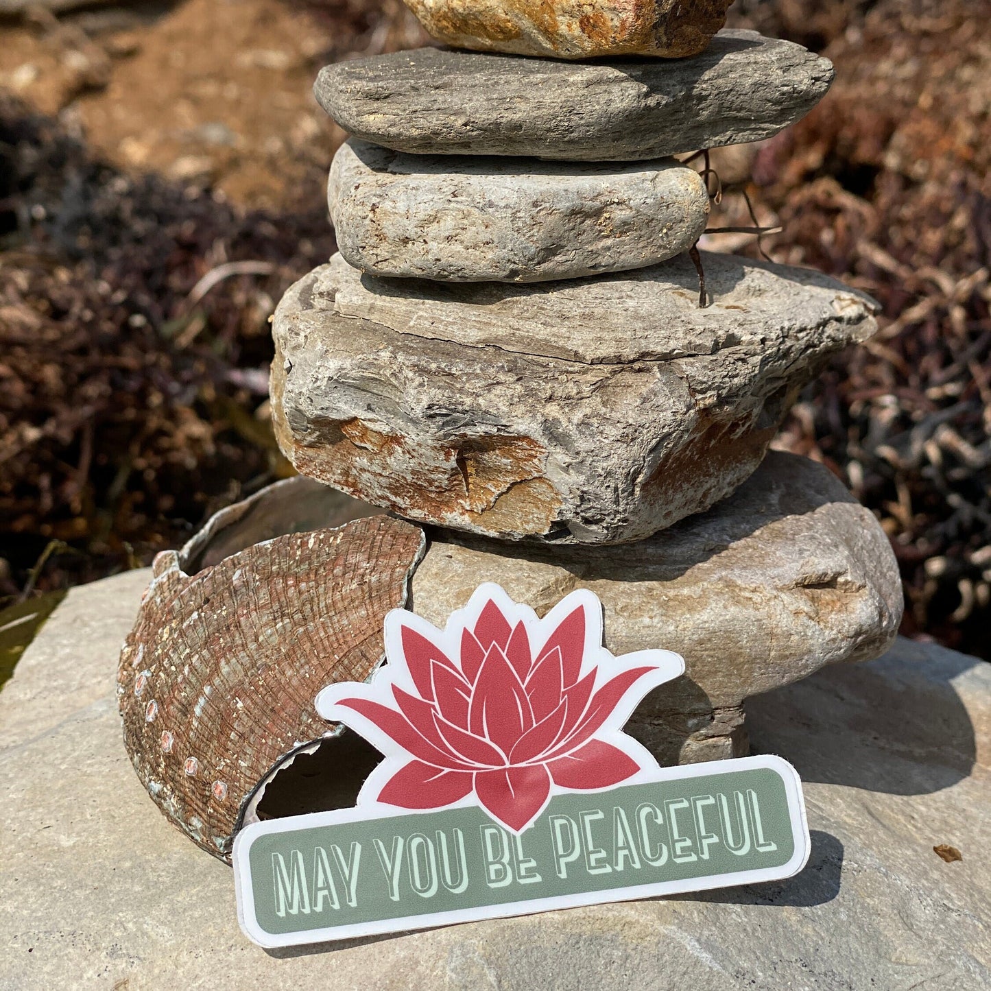 Stack of stones with a pink lotus flower sticker and 'May You Be Peaceful' text on a natural background.