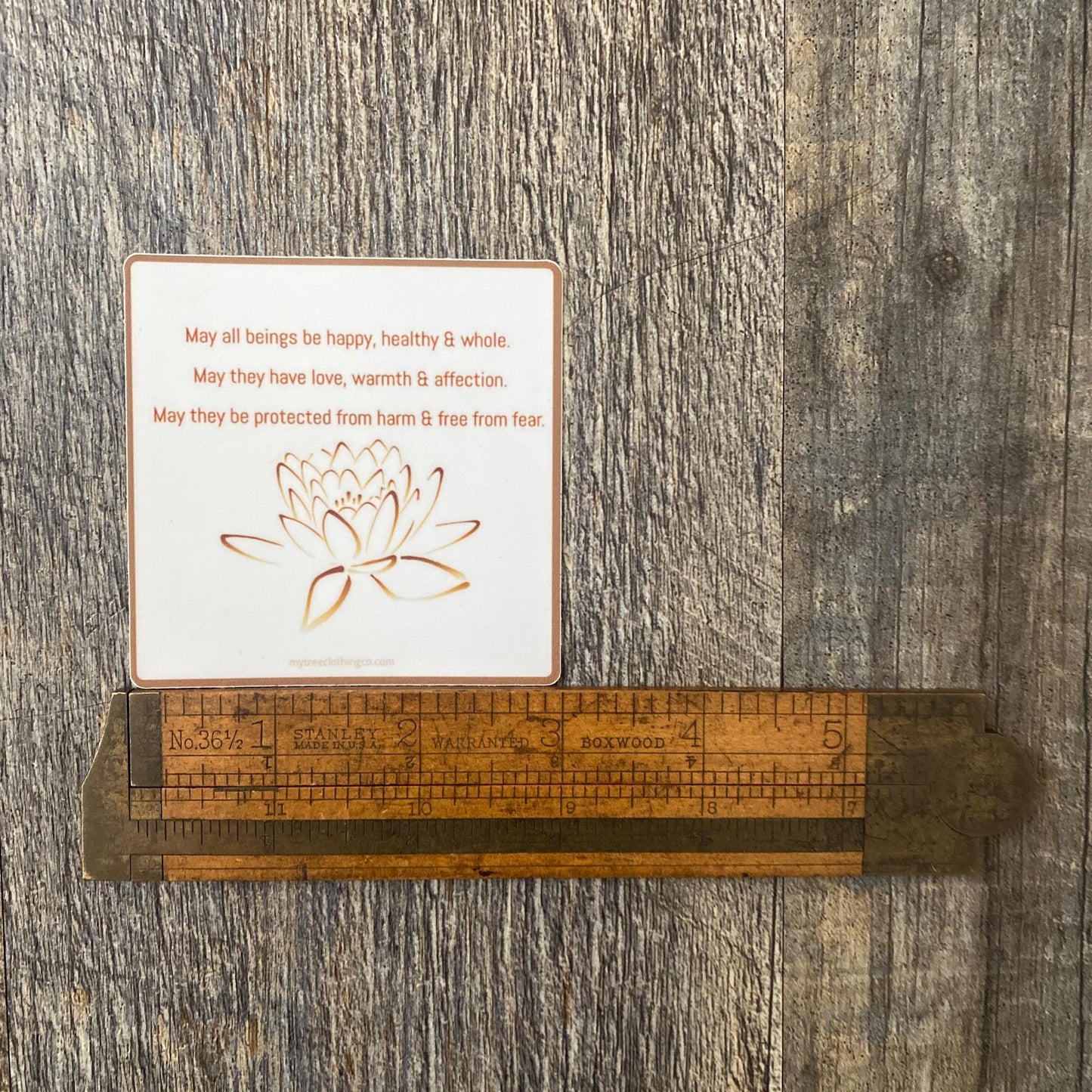3" square Buddhist sticker with a lotus flower design and text, 'May all beings be happy, healthy and whole. May they have love, warmth and affection. May they be protected from harm and free from fear.' On a wooden surface with a ruler for scale.