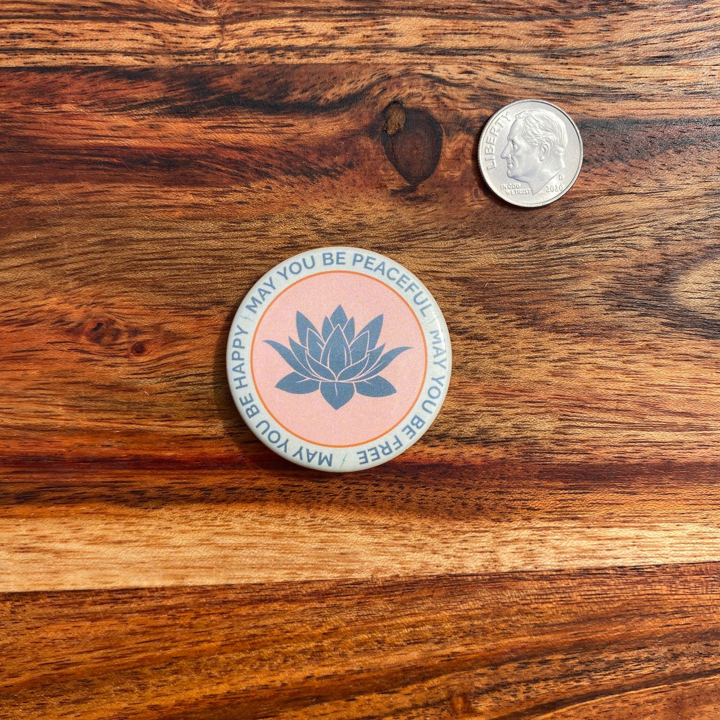 Circular sticker with lotus design and the text, may you be peaceful may you be free may you be happy, on a wooden surface next to a coin.