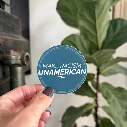 Hand holding a blue sticker with 'MAKE RACISM UNAMERICAN' text against a plant and industrial background.