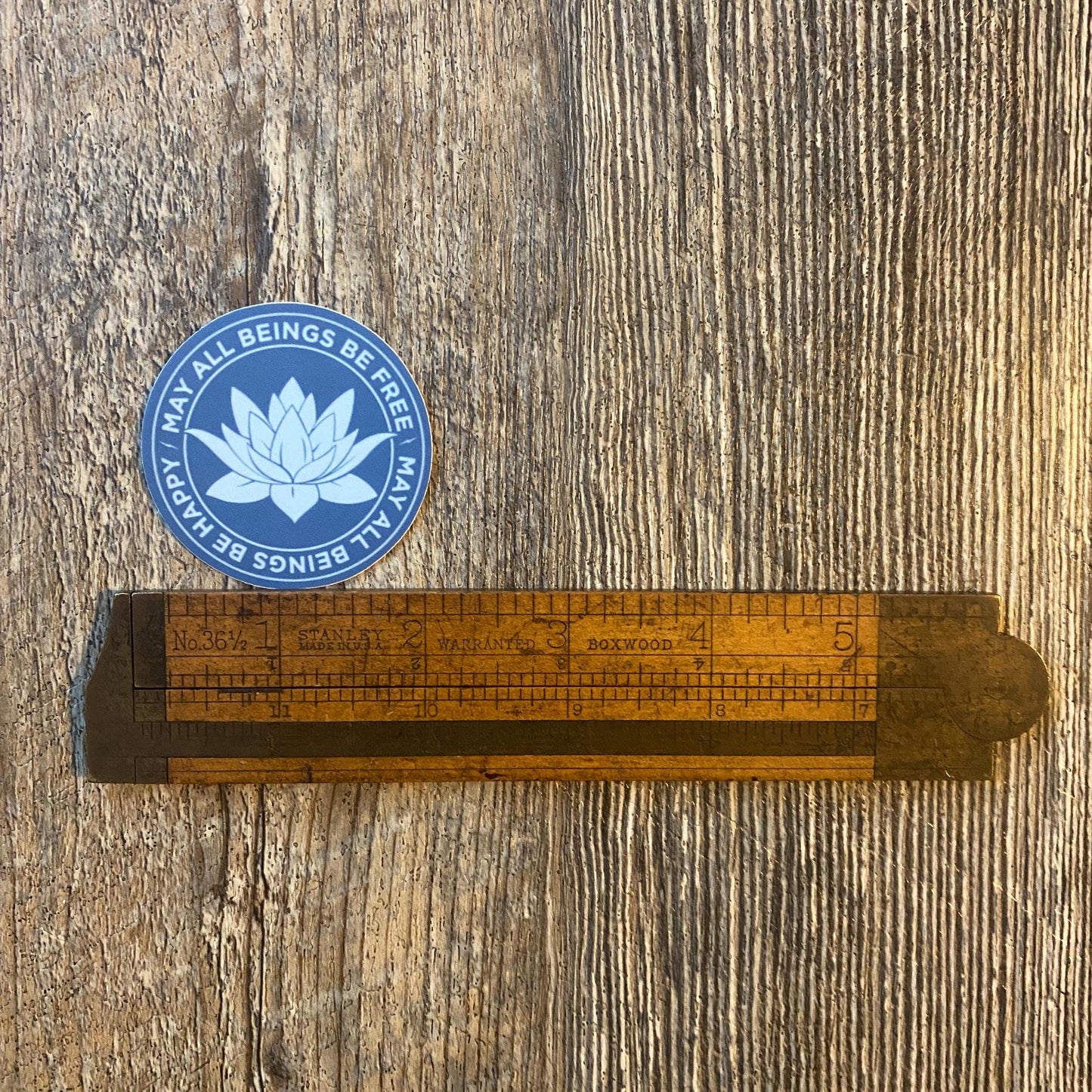Buddhist meditation sticker in a gorgeous blue with a lotus flower design and the text, 'may all beings be free may all beings be happy' on a wooden surface.