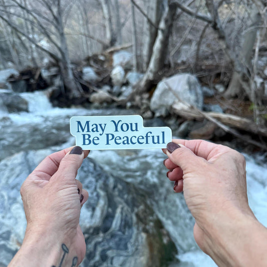 Hands holding light blue and dark blue 'May You Be Peaceful' sticker against a natural background of rocks and water.