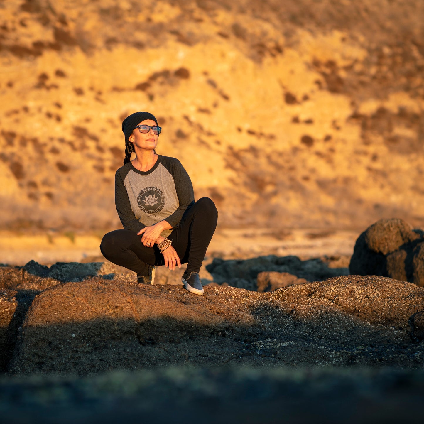 Person sitting on a rocky beach at sunset wearing a Gray and black 1/4 length sleeve shirt with a lotus logo, and the text, may all beings be free may all beings be happy.