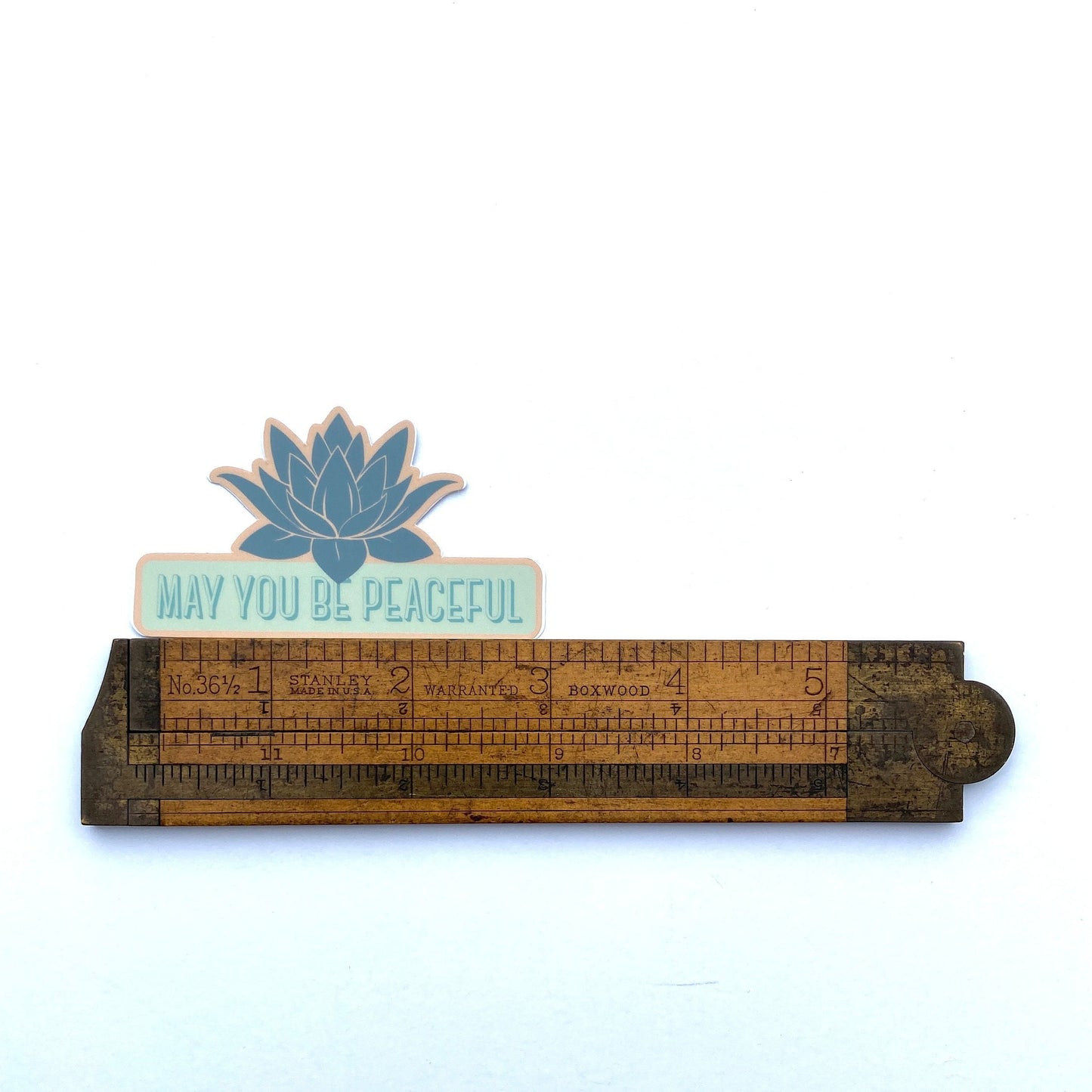 Wooden ruler with a blue lotus flower sticker and the text, 'May You Be Peaceful' on a white background.