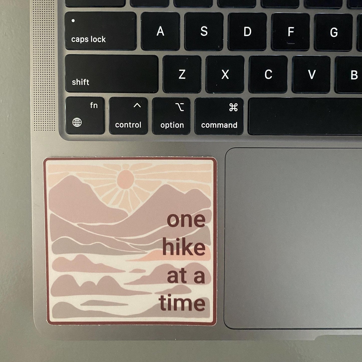 3" square sticker with 'one hike at a time' in neutral colors with mountains and a sunset on the keyboard.