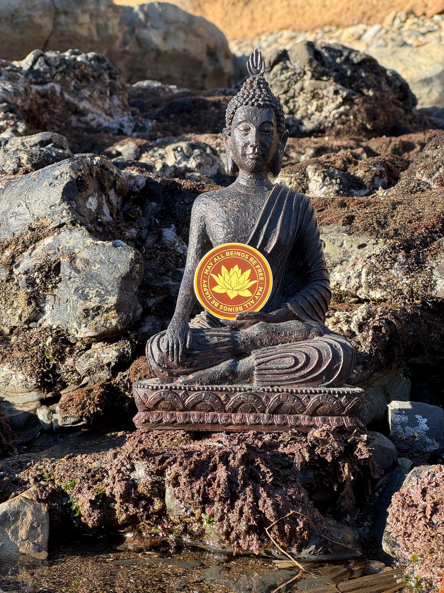 Buddha statue on rocks with a 2" round saffron and gold vinyl sticker with Buddhist mettā prayer text, may all beings be free, may all being be happy.