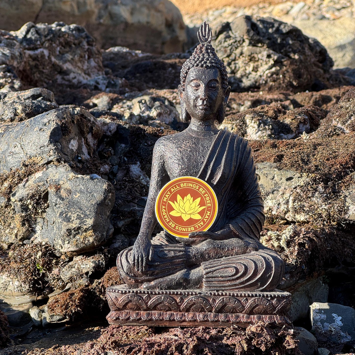 Buddha statue on rocks with a 2" round saffron and gold vinyl sticker with Buddhist mettā prayer text, may all beings be free, may all being be happy.
