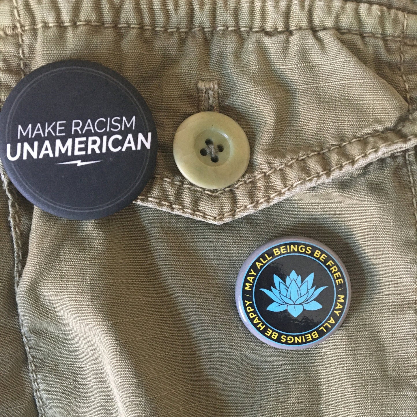Two buttons on a khaki background, one with 'Make Racism Unamerican' and one 1.25" black and blue lotus button with the Buddhist prayer, may all beings be free may all beings be happy.