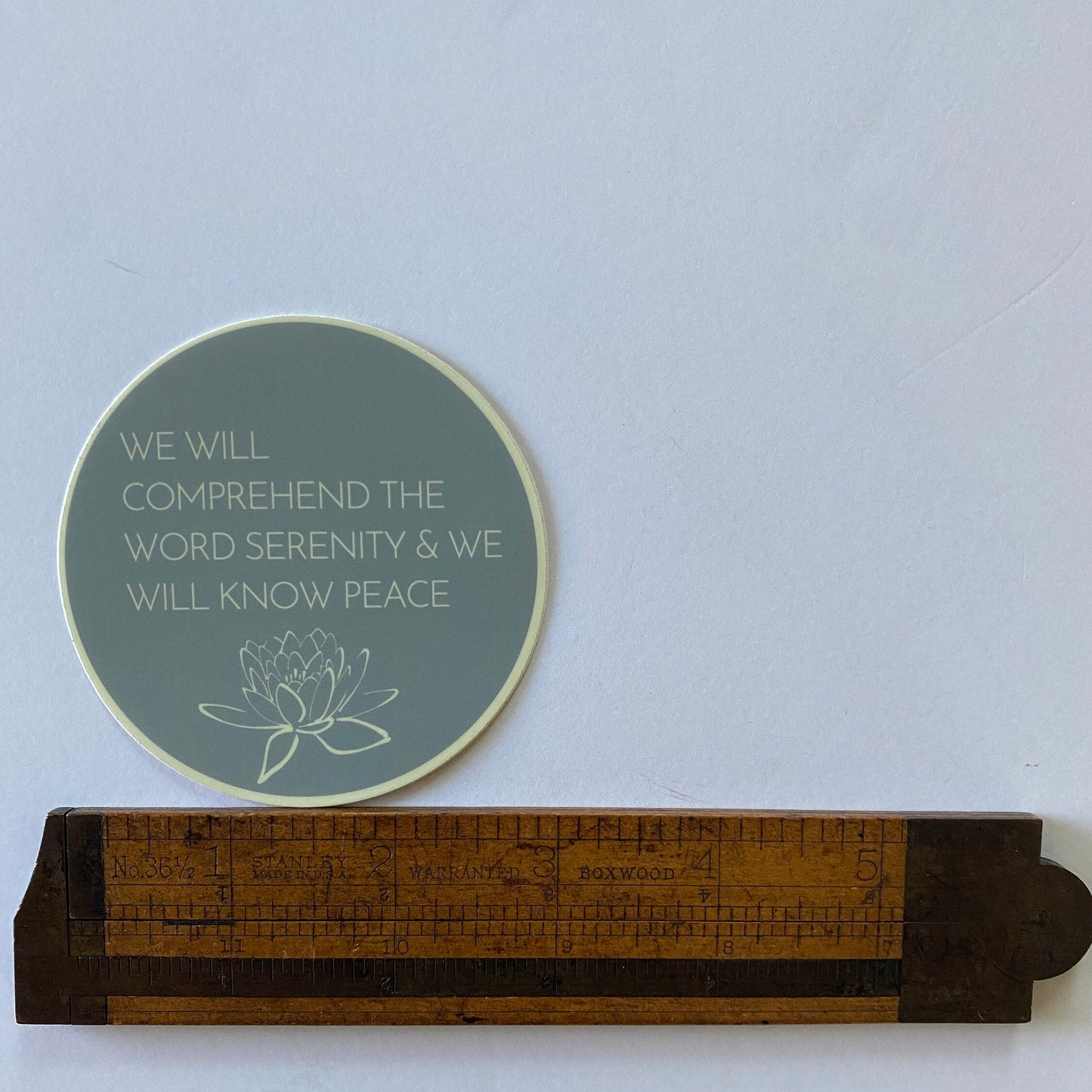 Round teal sticker with, WE WILL COMPREHEND THE WORD SERENITY AND WE WILL KNOW PEACE, 12 step recovery quote design next to a wooden ruler on a light blue background