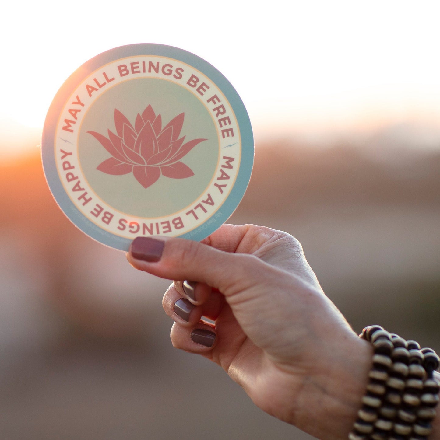Hand holding a 4" circle lotus flower design and text, MAY ALL BEINGS BE FREE MAY ALL BEINGS BE HAPPY, against a blurred background.