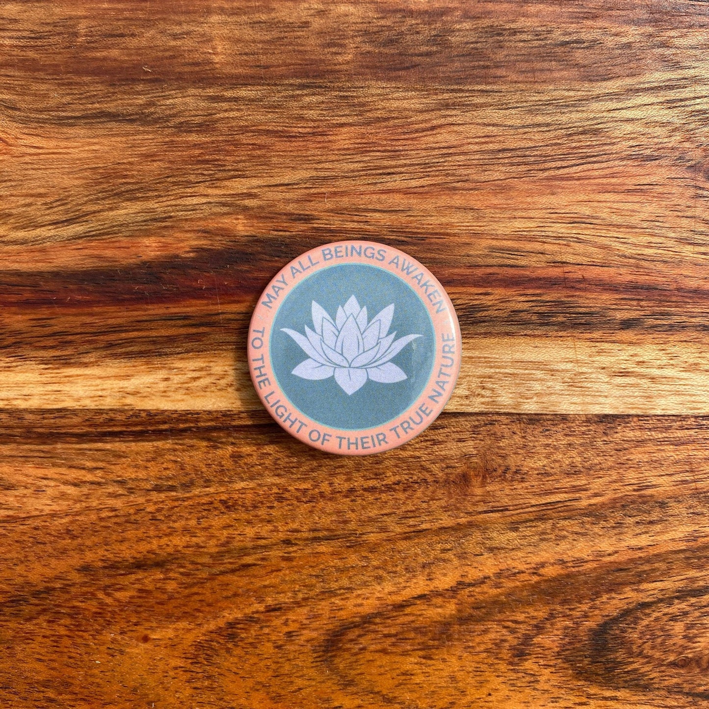 Round button with lotus flower design and text, may all beings awaken to the light of their true nature, on a wooden surface.