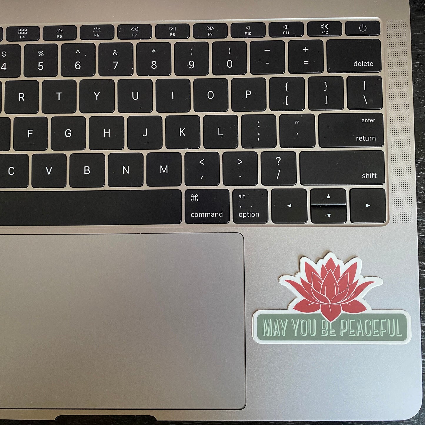 Laptop keyboard with a red lotus 'May You Be Peaceful' sticker on a metallic surface.