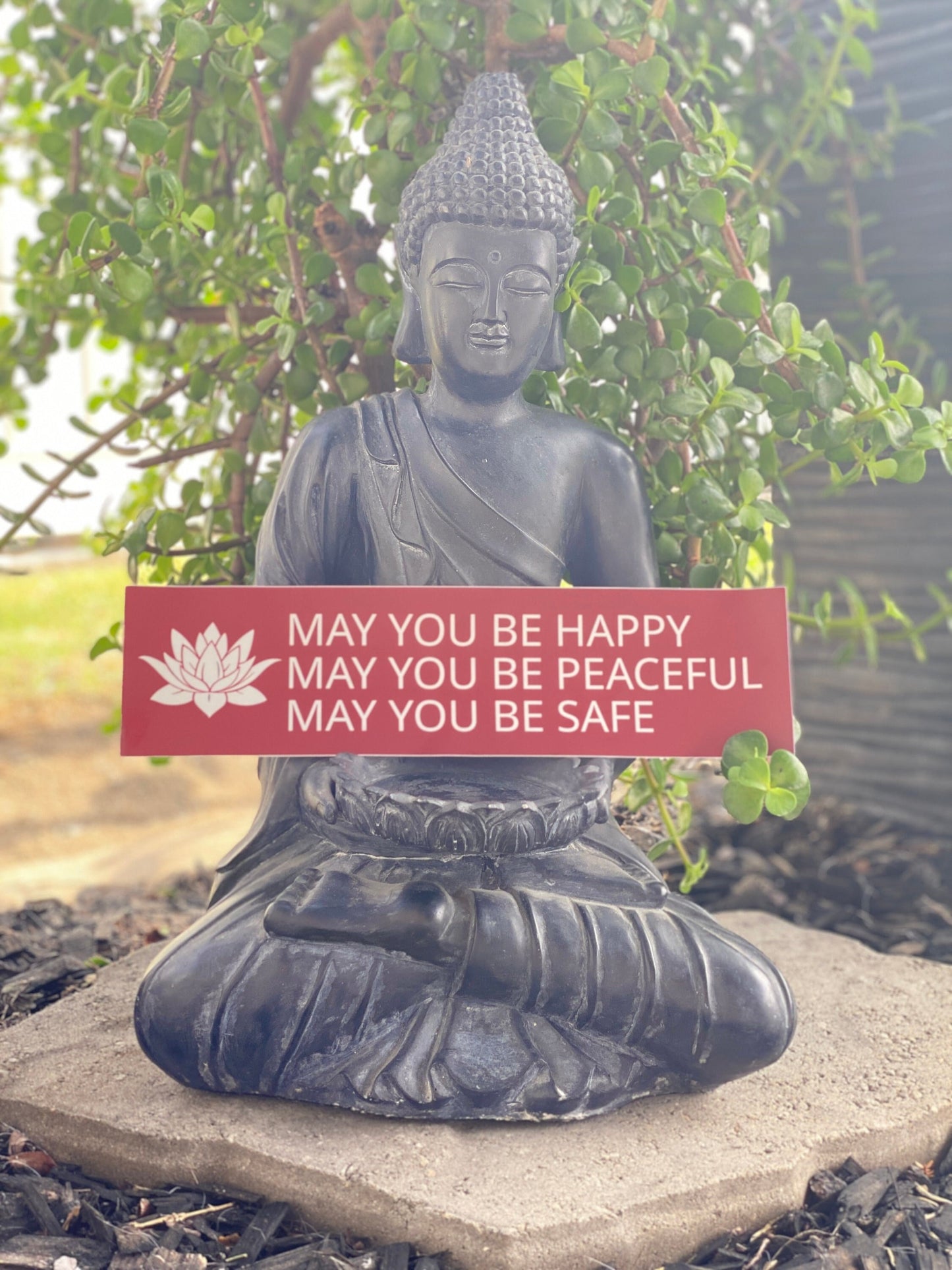 Buddha statue with a Red and white bumper sticker with a lotus and the prayer, may you be happy may you be peaceful may you be safe in a garden setting.
