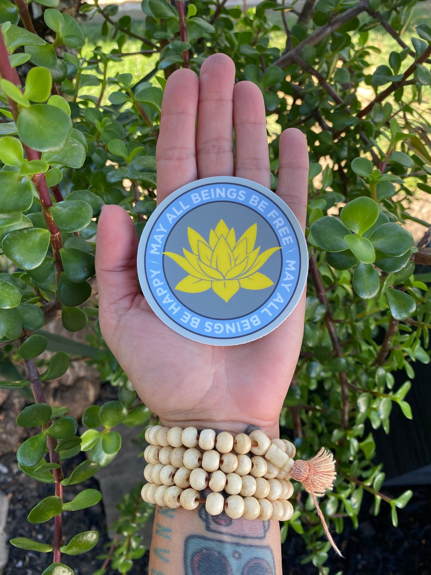 Hand holding a 3" circular sticker with a lotus flower design and text, may all beings be free may all beings be happy, against a green leafy background.