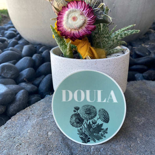 Round 'DOULA' sticker with floral design on a stone surface next to a potted plant.