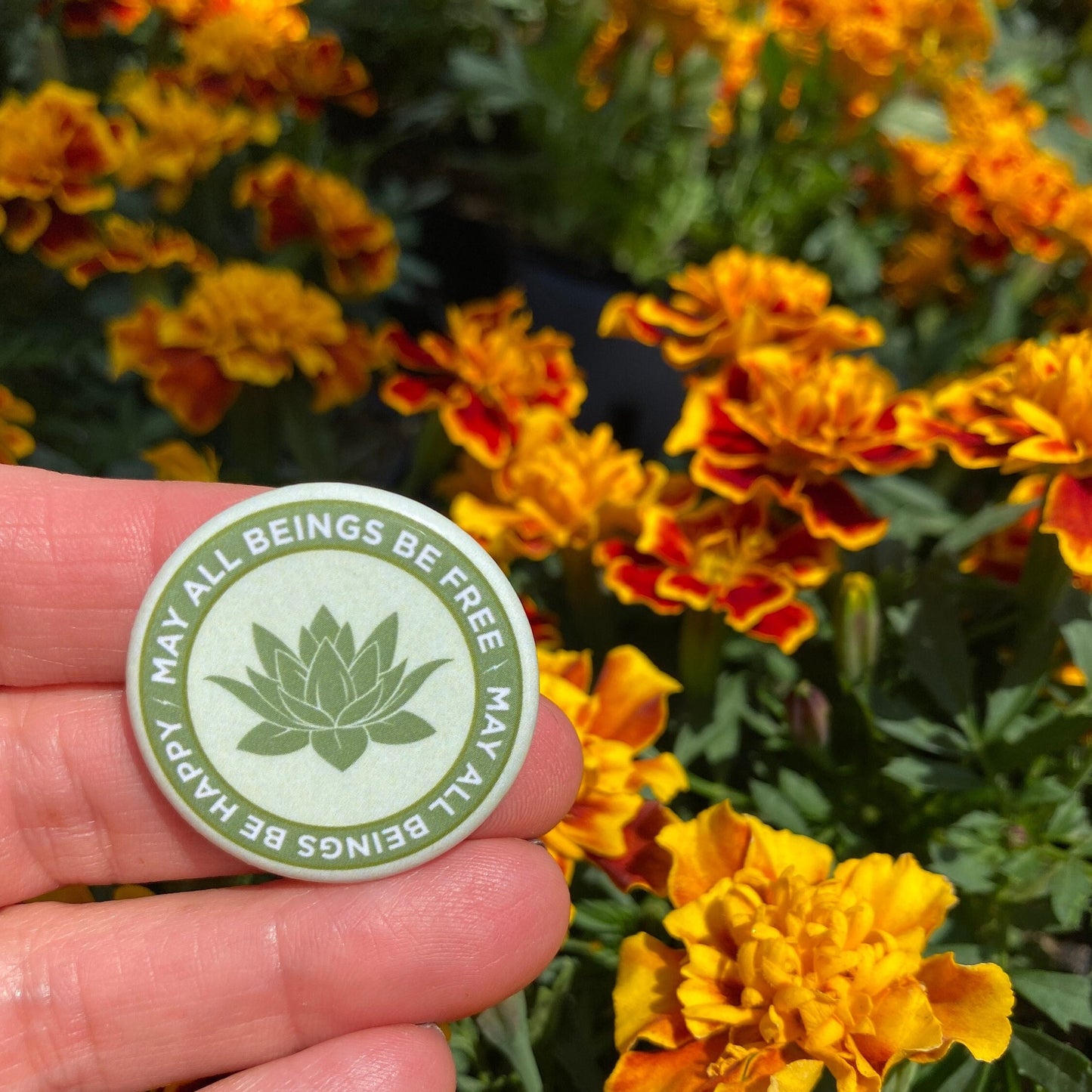 Hand holding a light green 1.25" circular Buddhist pin back button with text and a lotus symbol in front of marigold flowers.