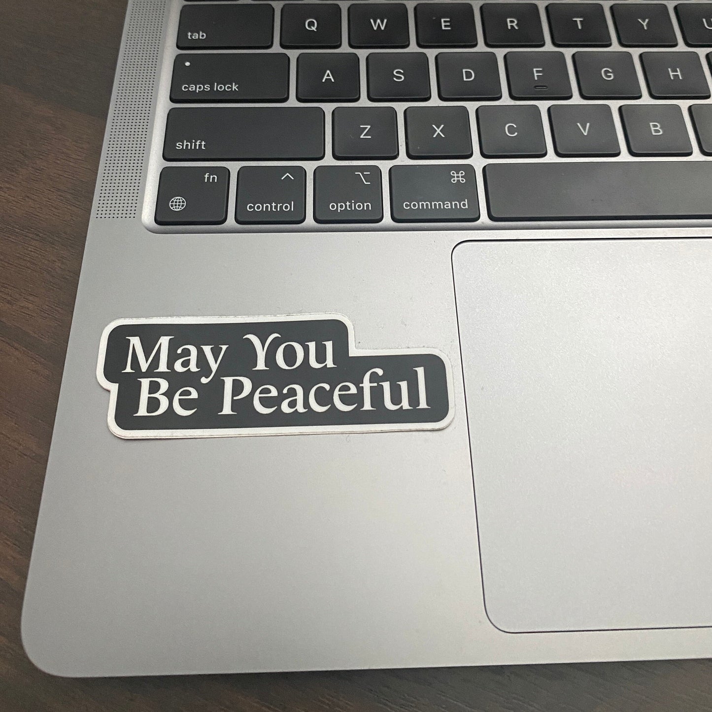Laptop with a sticker reading 'May You Be Peaceful' on a wooden surface