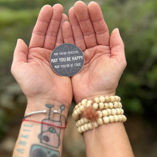 Two hands holding a 2" circular sticker with text, may you be peaceful may you be happy may you be at ease, against a blurred natural background.