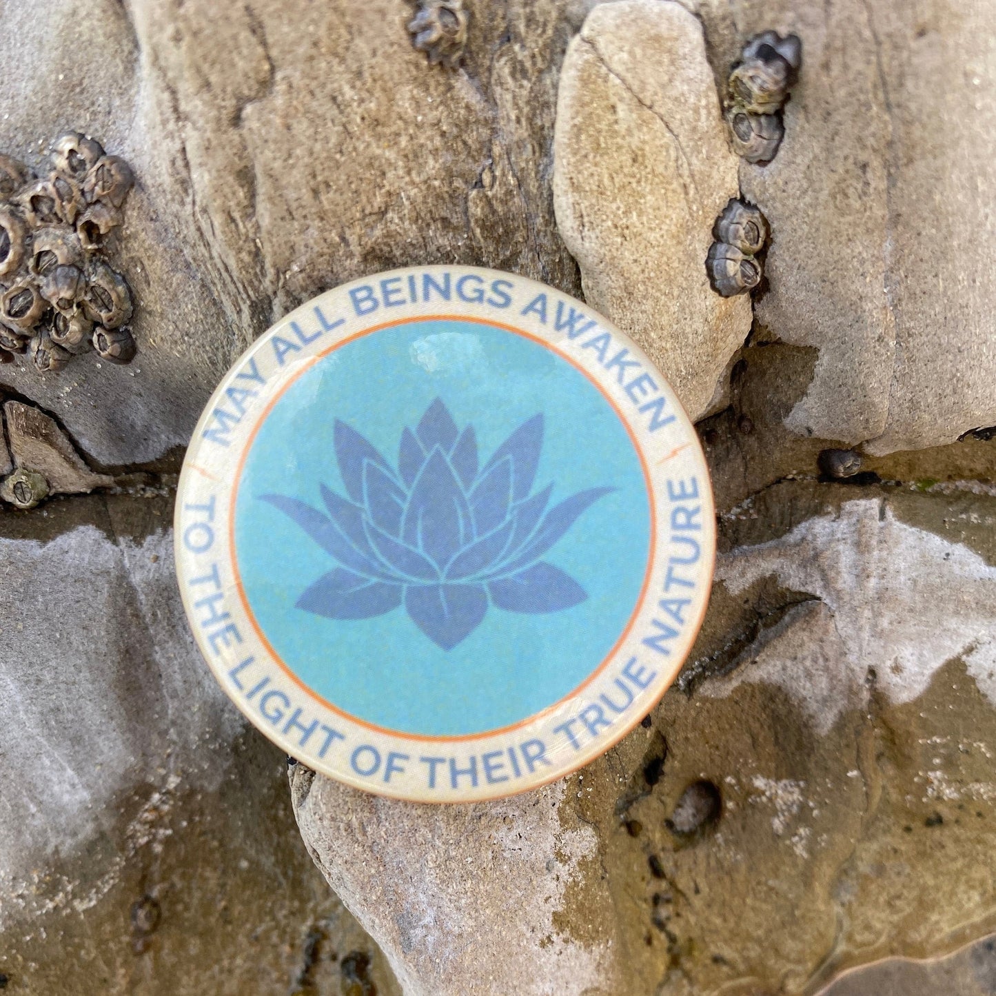1.5" button with the mantra that reads, may all beings awaken to the light of their true nature, with a lotus design on a stone surface.