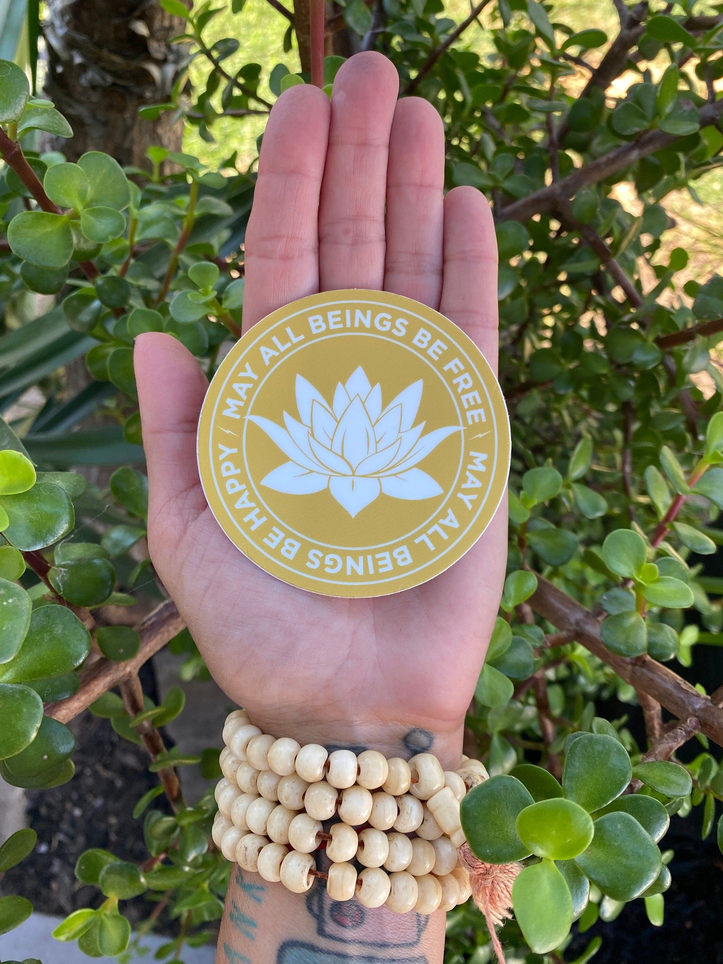 Hand holding a 3" gold and white sticker with a lotus flower design and text, may all beings be free may all beings be happy, against a green leafy background.