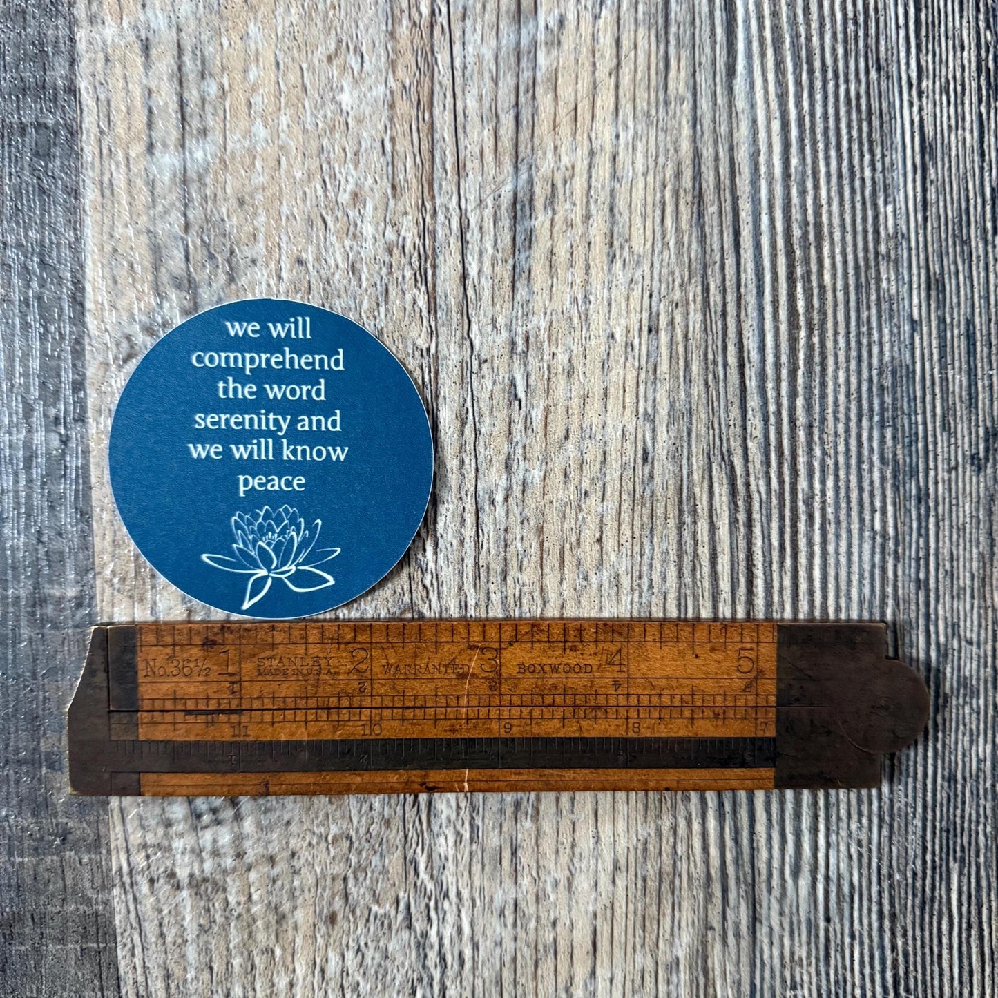2.5" blue sticker with a quote from the big book of AA, "we will comprehend the word serenity and we will know peace" and a lotus flower on a wooden ruler against a wooden background.