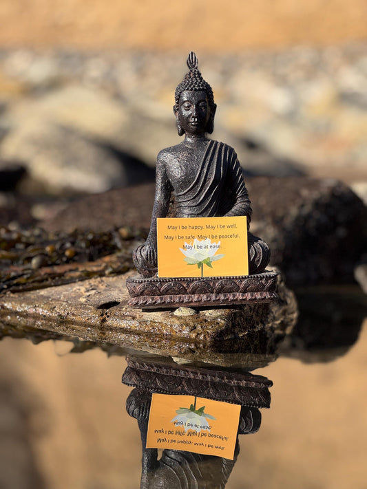 Small Buddha statue with a rectangular yellow sticker with a lotus and the words, may i be happy may i be well may i be safe may i be peaceful on a reflective surface.