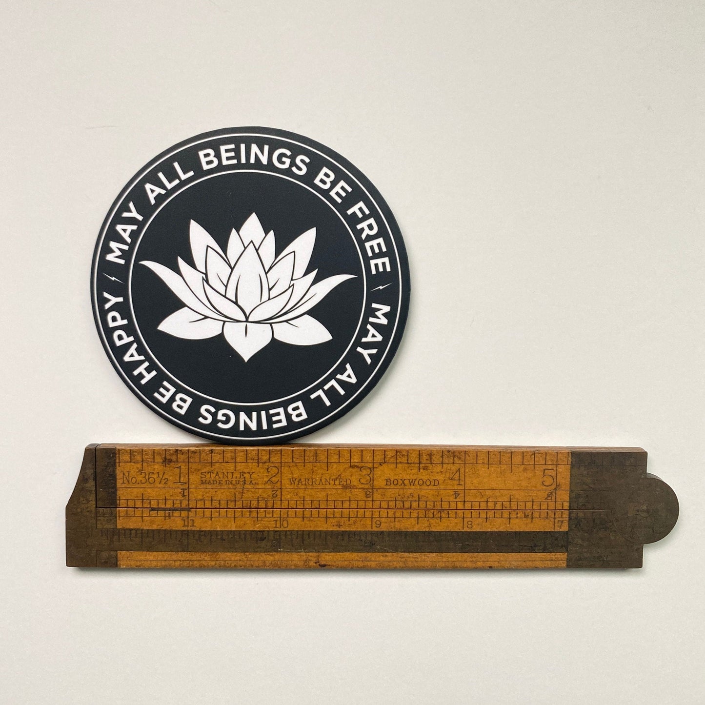3.5" round black and white magnet with lotus flower design and text, may all beings be free, may all beings be happy. It is being measured by a wooden ruler.