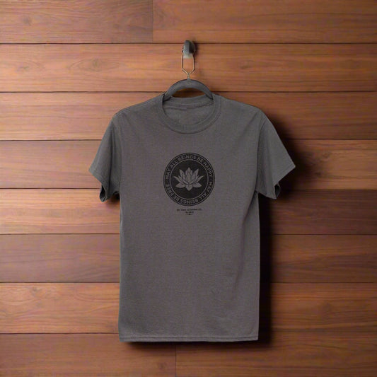 Gray t-shirt with a black logo on a hanger against a wooden background. The shirt has a lotus in the center and the text, may all beings be happy may all beings free from the Buddhist loving kindness prayer.