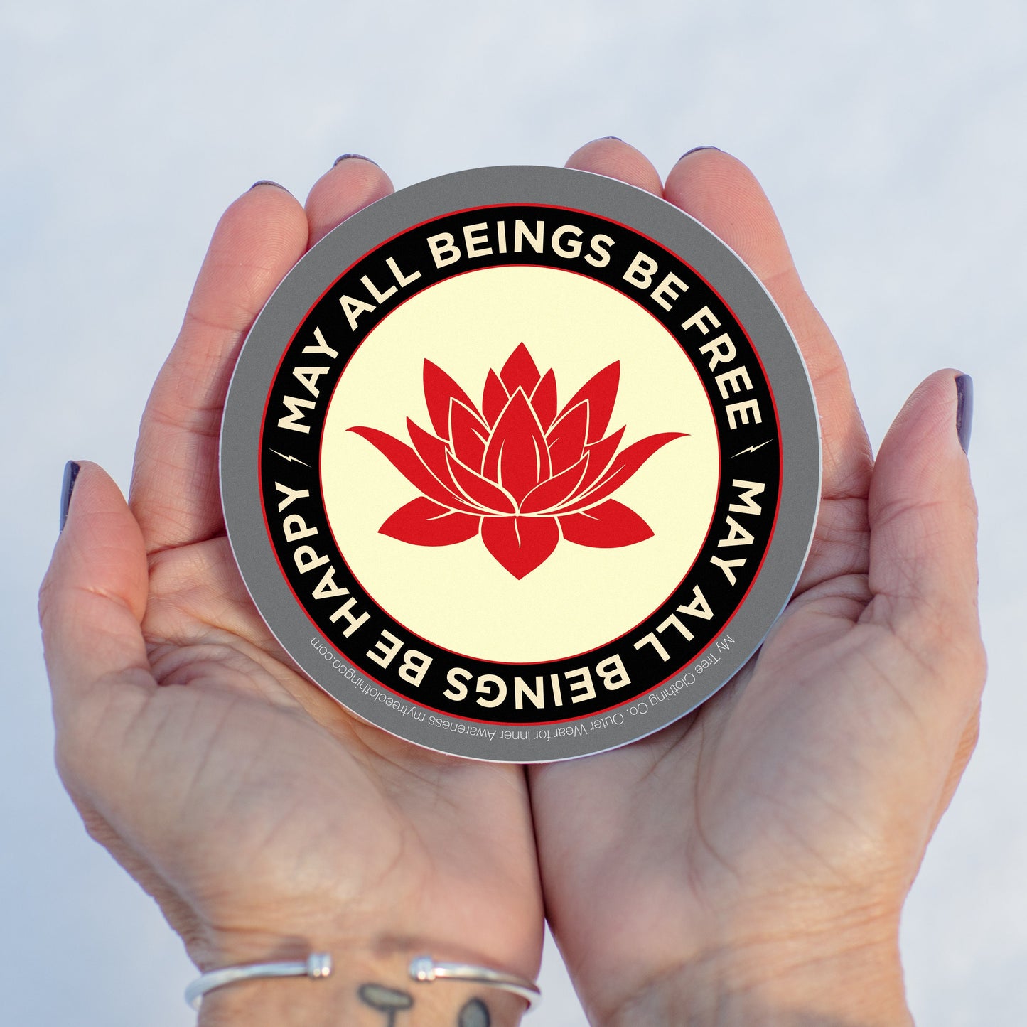 Hand holding a circular sticker with a red lotus flower and text may all beings be free may all beings be happy, against a light background.