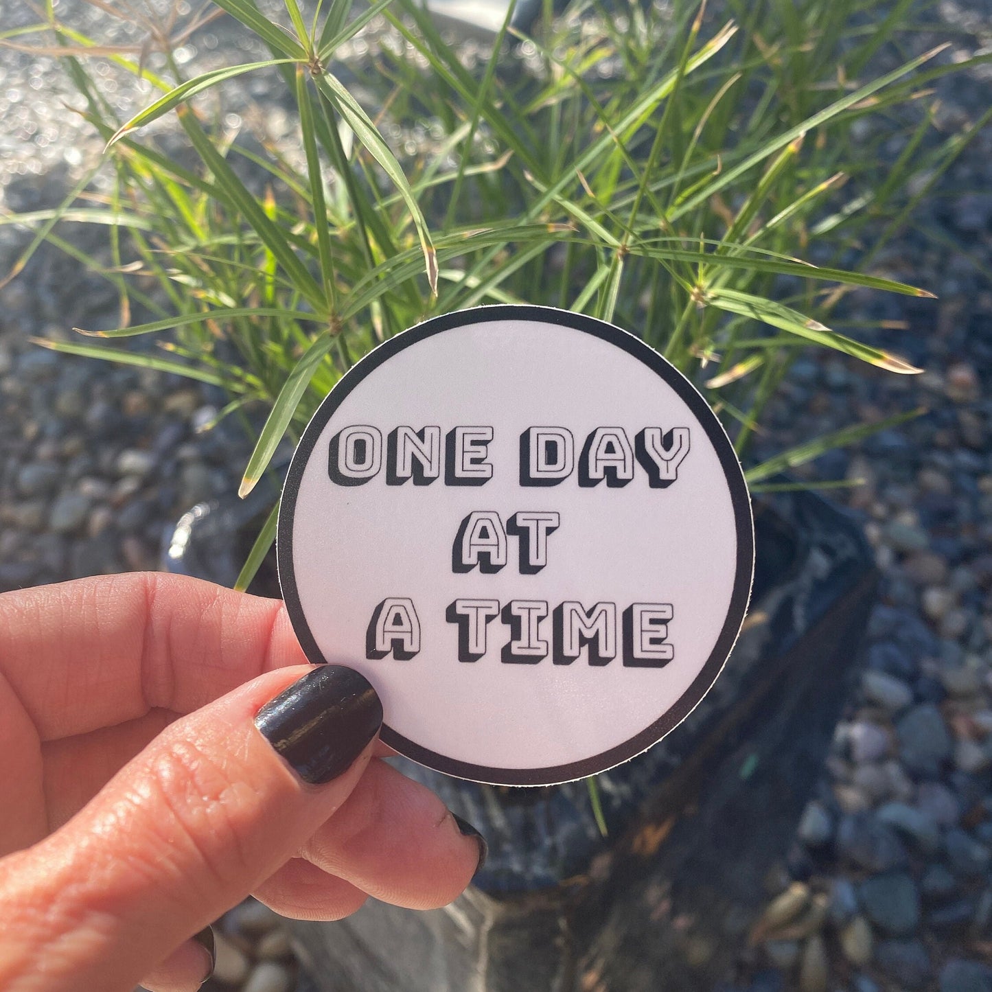 Hand holding a 'One Day at a Time' sticker with greenery in the background
