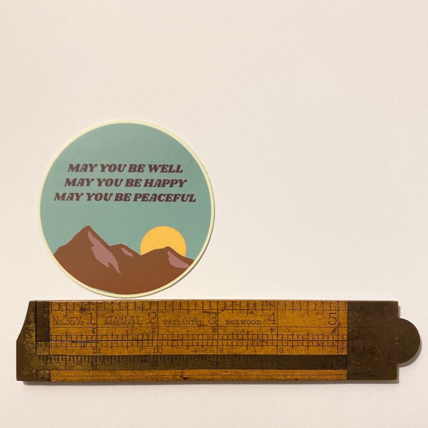 Decorative ruler with a circular Buddhist sticker design featuring mountains and text, may you be well may you be happy may you be peaceful, on a beige background.