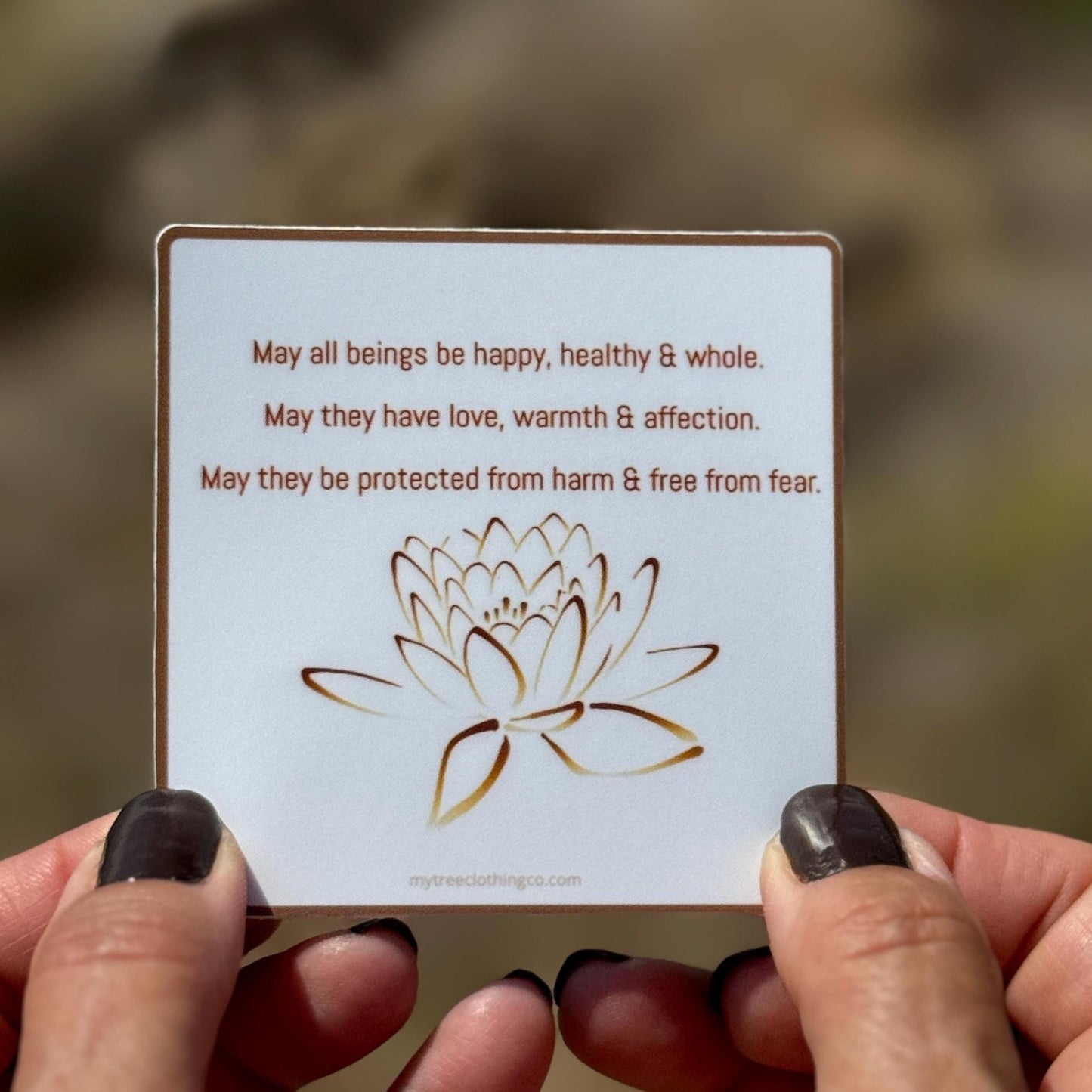 3" square Buddhist sticker with a lotus flower design and text, 'May all beings be happy, healthy and whole. May they have love, warmth and affection. May they be protected from harm and free from fear.' Held by hands against a blurred natural background.