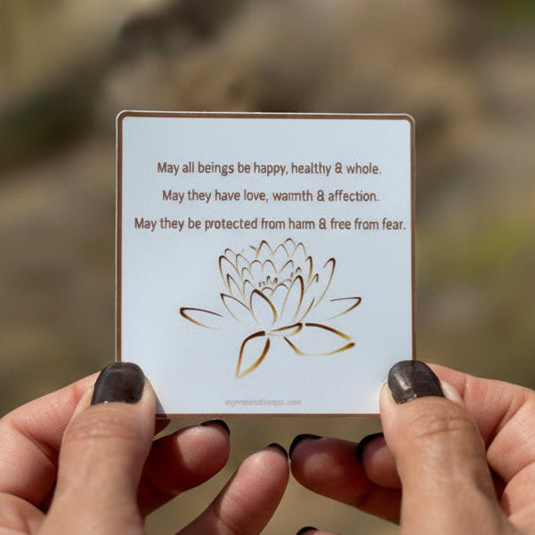 3" square Buddhist sticker with a lotus flower design and text, 'May all beings be happy, healthy and whole. May they have love, warmth and affection. May they be protected from harm and free from fear.' Held by hands against a blurred natural background.