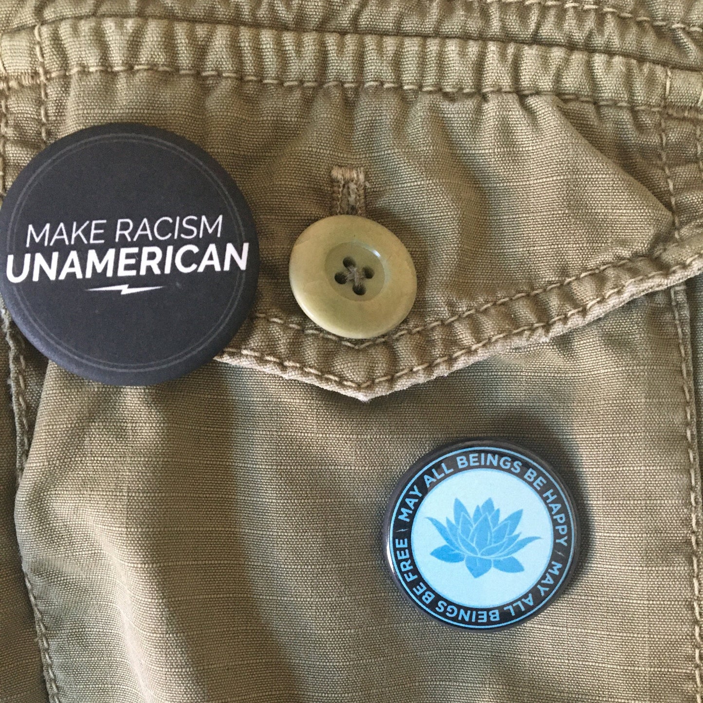 Button with 'Make Racism Unamerican' text on a beige jacket with another, circular blue and black 1.25" button with a lotus flower design and text, may all beings be happy may all beings be free.