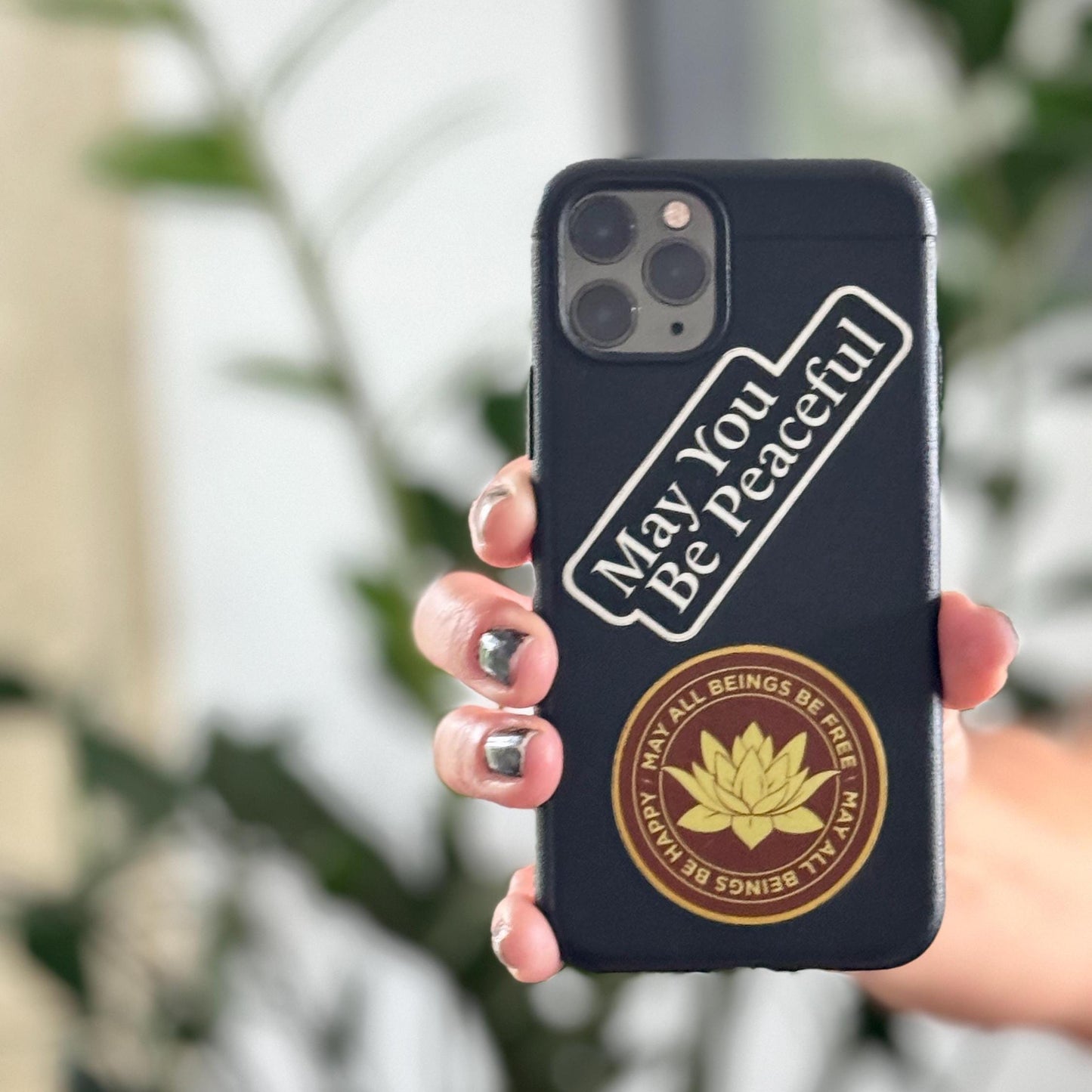 Phone case with 'May You Be Peaceful' text and lotus flower design held by a hand against a blurred natural background.